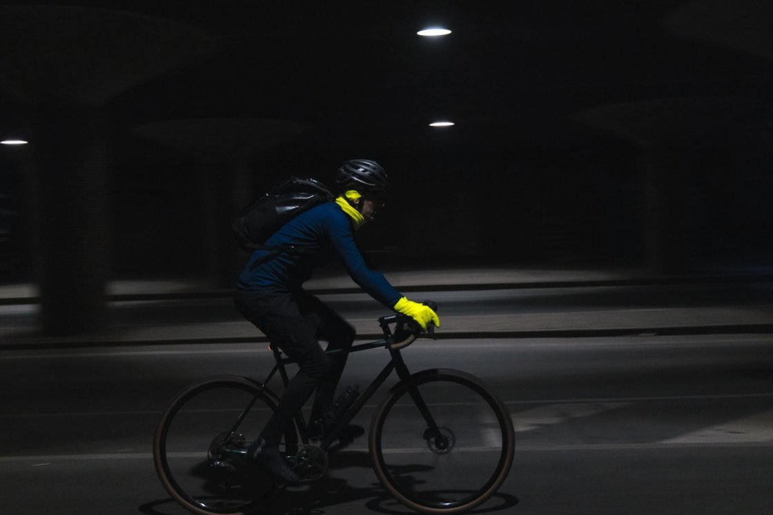 Eine Person auf einem dunklen Rennrad fährt quer zur Blickrichtung nach rechts. In der nächtlichen Szenerie sind lediglich die neongelben Handschuhe und ein neongelbes Halstuch klar erkennbar.