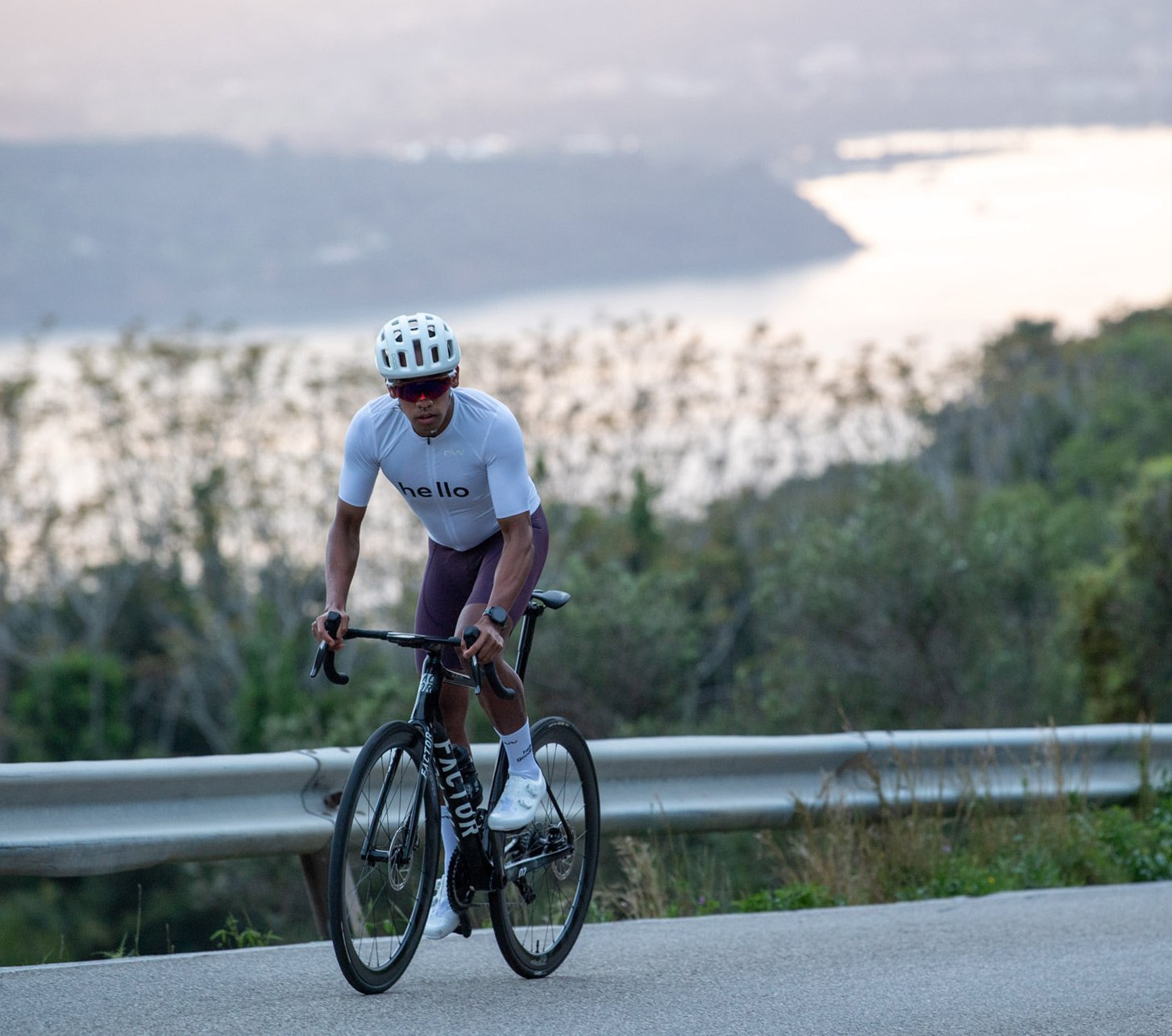 Ein Rennradfahrer, der Northwave Rennrad Bekleidung und Northwave Schuhe trägt, fährt an einem sonnigen Tag im Wiegetritt eine asphaltierte Straße mit leichter Steigung hoch. Im Hintergrund ist ein See zu sehen. 