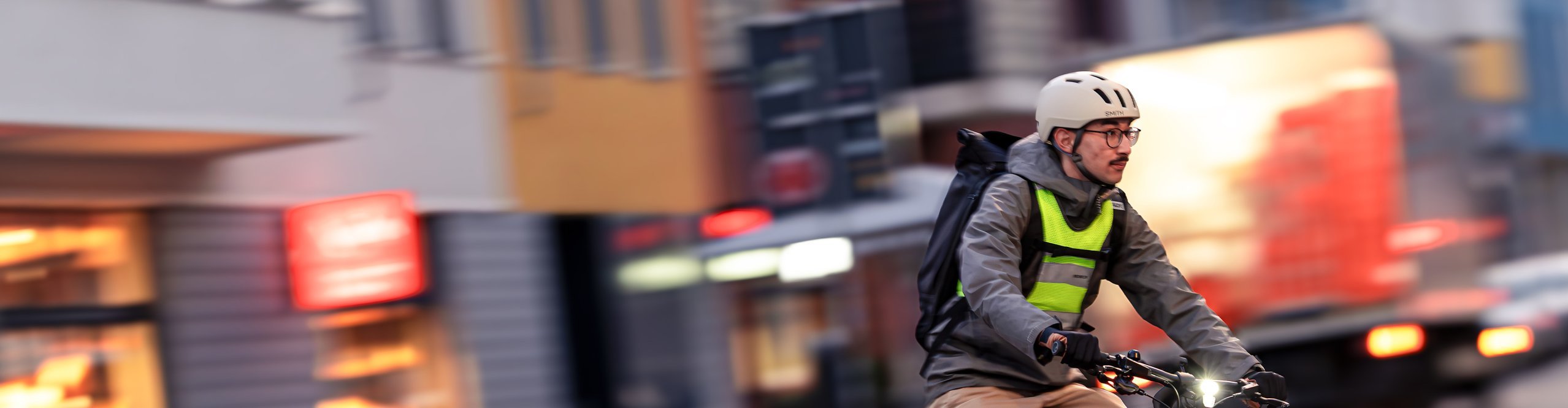 Pendler fährt mit E-Bike durch die Stadt, mit Helm, Rucksack und Beleuchtung im Straßenverkehr, nachhaltige Alternative bei hohen Benzinpreisen