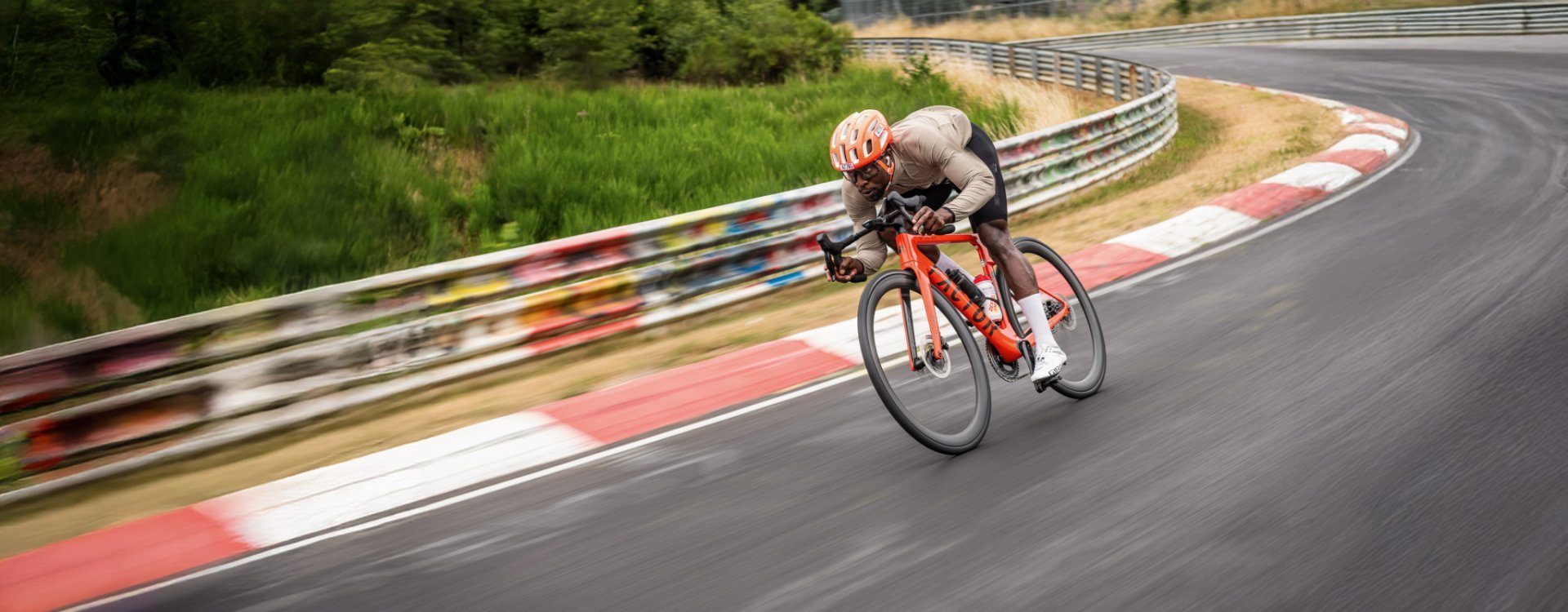 Ein sportlicher, junger Mann mit orangem Helm fetzt auf einem roten Rennrad über eine Auto-Rennstrecke 
