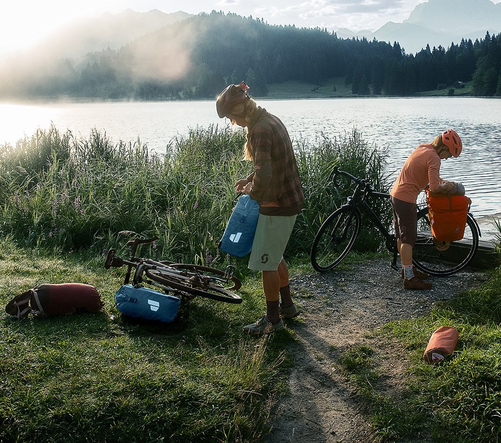 Bikepacking am See bei Sonnenaufgang: Zwei Radfahrer packen wasserdichte Deuter Fahrradtaschen und Gepäckbeutel neben ihren Gravelbikes in alpiner Natur.