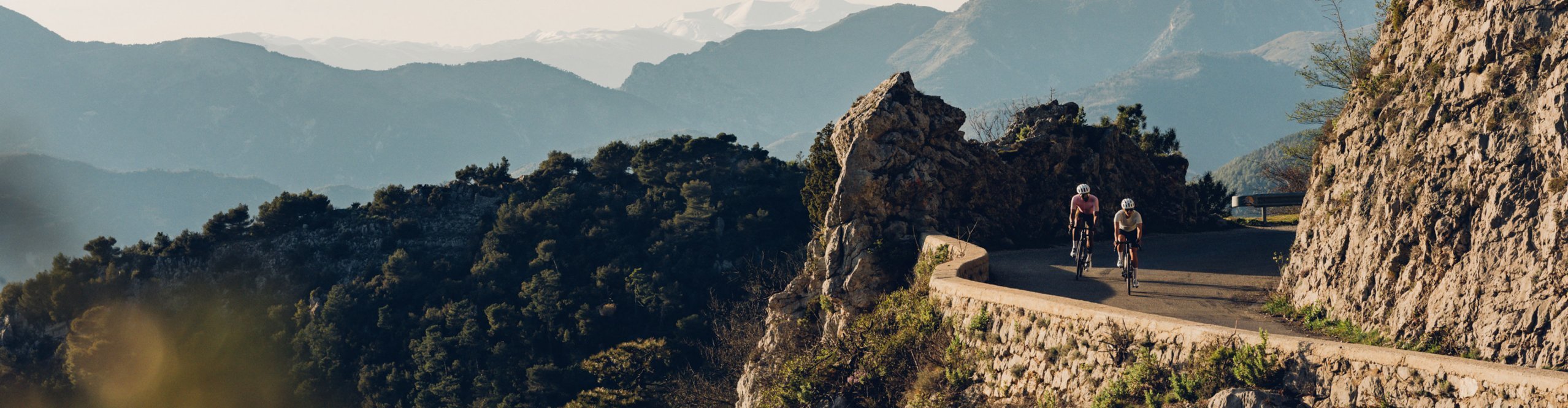 Zwei Rennradfahrer fahren auf kurviger Bergstraße zwischen Felsen durch die Alpenlandschaft – sportlicher Aufstieg in beeindruckender Kulisse bei tiefstehender Sonne