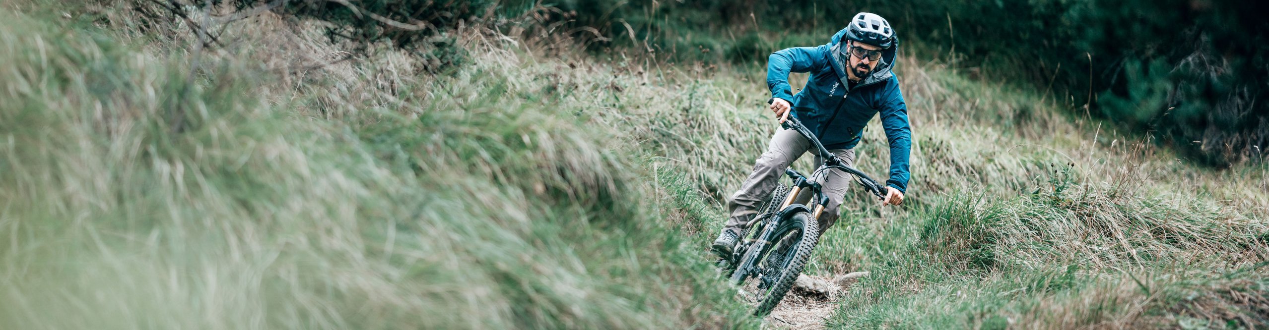 Mountain biker wearing a blue Dirtlej jacket and grey MTB trousers on a narrow trail through a wooded hillside.