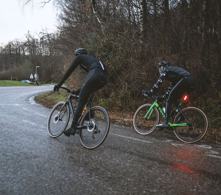 Stephan et Sebastian, du service clientèle de bc, roulent sur une route mouillée par la pluie avec leurs vélos de route. 