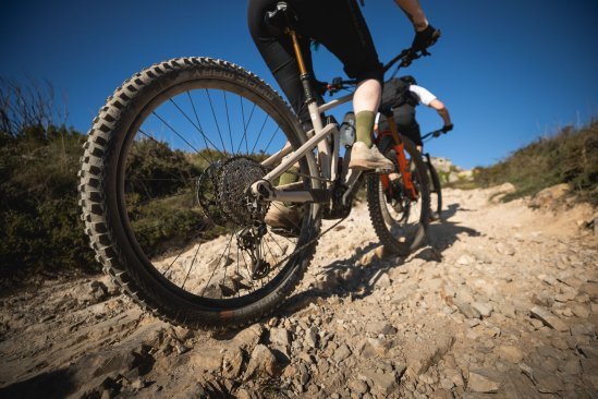 Zwei E-MTB kraxeln einen felsigen Trail hinauf. Die Personen sind star kangeschnitten, Fokus liegt auf dem Hinterrad und Antrieb des hinteren Bikes. Der Himmel ist kräftig blau.