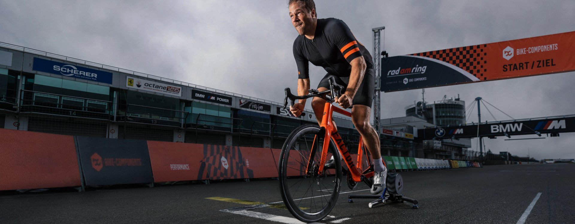 A man on an orange road bike trains on a smart trainer at the start line of the Nürburgring racetrack during the “Rad am Ring” event.