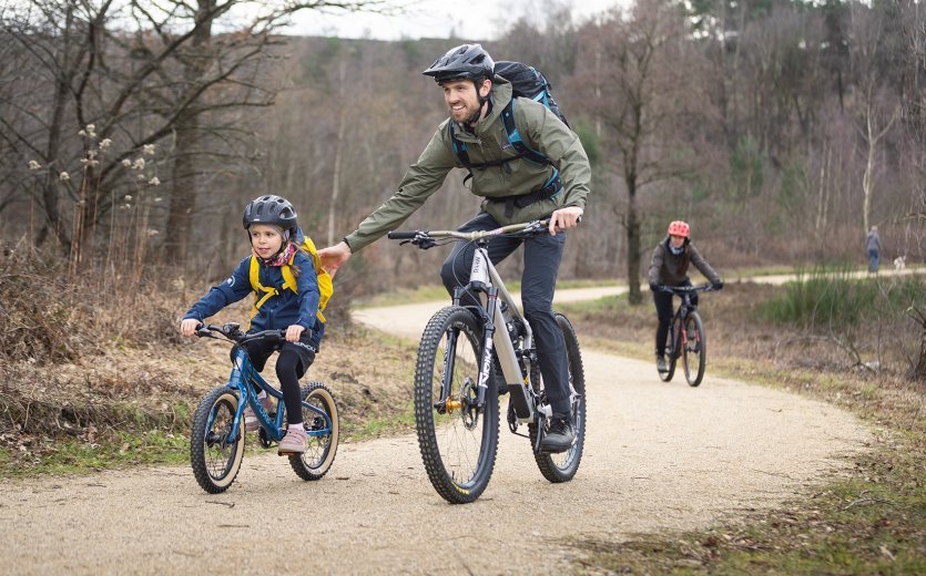 Ein Kind fährt mit einem Erwachsenen Mountainbike. Sie sind auf einem Schotterweg unterwegs.