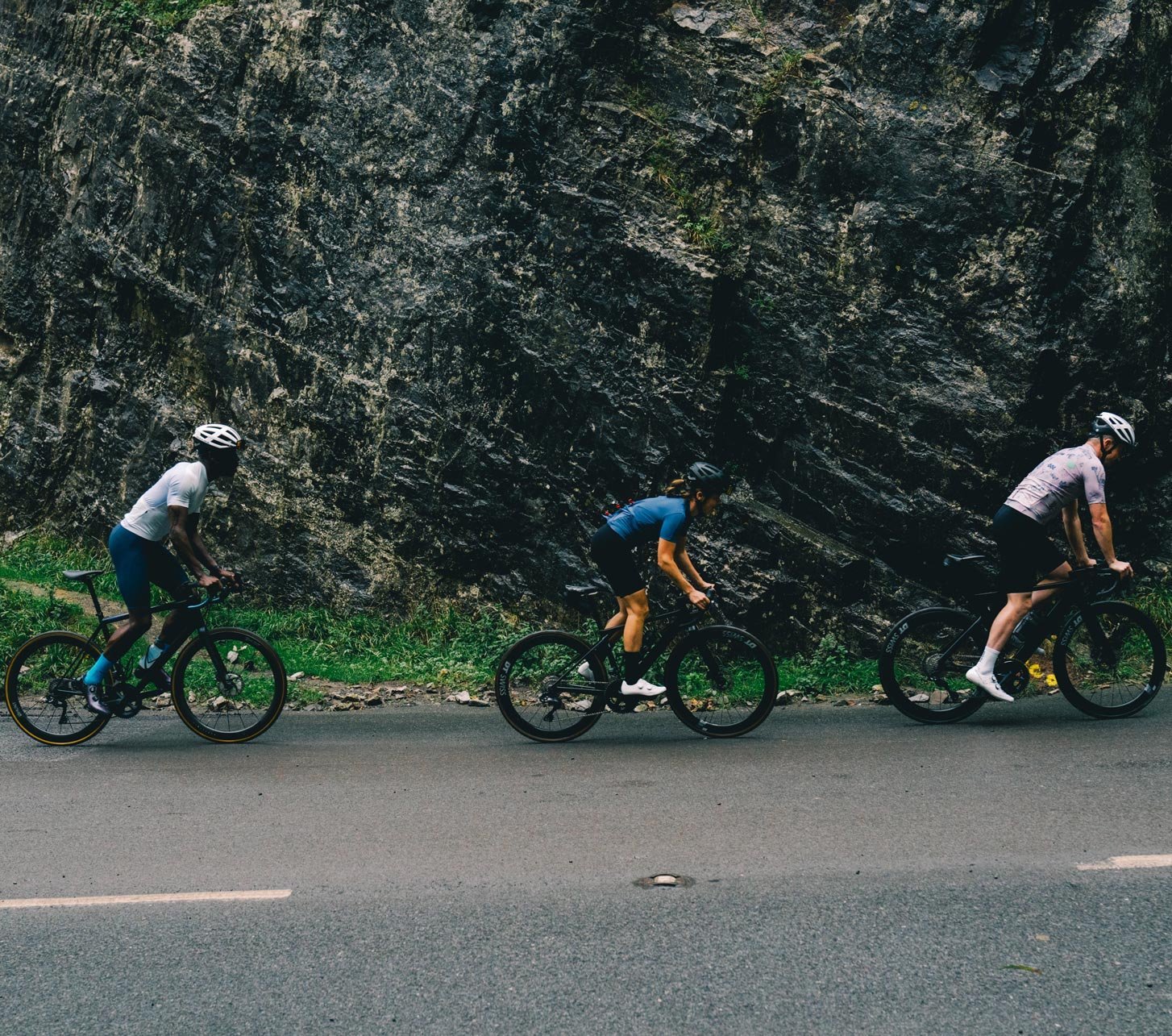 Tres ciclistas de carretera con ropa de ciclismo Endura pedaleando por una carretera de montaña junto a una pared rocosa