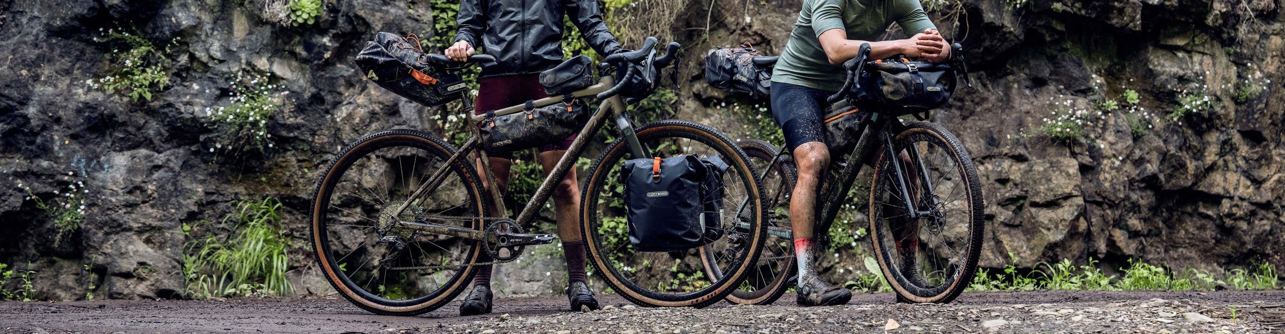 Two bikepackers with gravel bikes and ORTLIEB bikepacking bags on a gravel trail in front of a rocky cliff