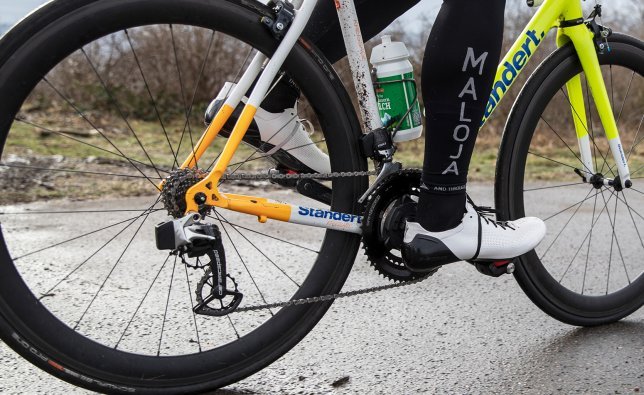 A road cyclist has optimised his bike with CeramicSpeed derailleur pulleys. He rides on a wet street.