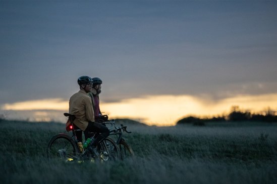 Dos ciclistas con cascos están con sus bicicletas en un campo al atardecer. Sus bicicletas tienen luces, el cielo es azul oscuro con un horizonte naranja y los árboles son visibles al fondo.