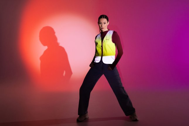 Eine junge Frau posiert vor einem bunt orange und pink angestrahlten Papierhintergrund. Zu einer schwarzen Hose trägt sie eine Leuchtweste von Wowow