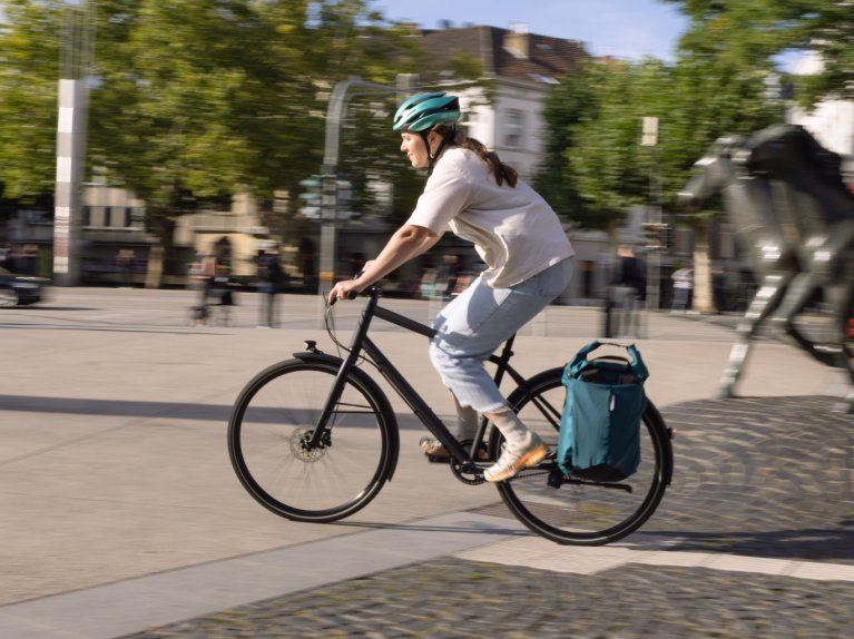A person wearing a helmet rides a bicycle across an urban square; in the background, pedestrians and several horse sculptures can be seen.