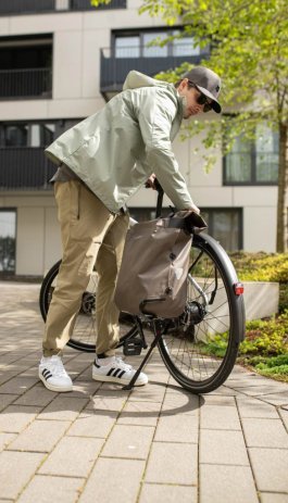 Une personne fixe un sac à vélo étanche Ortlieb au porte-bagages d'un vélo de ville devant un immeuble résidentiel moderne.