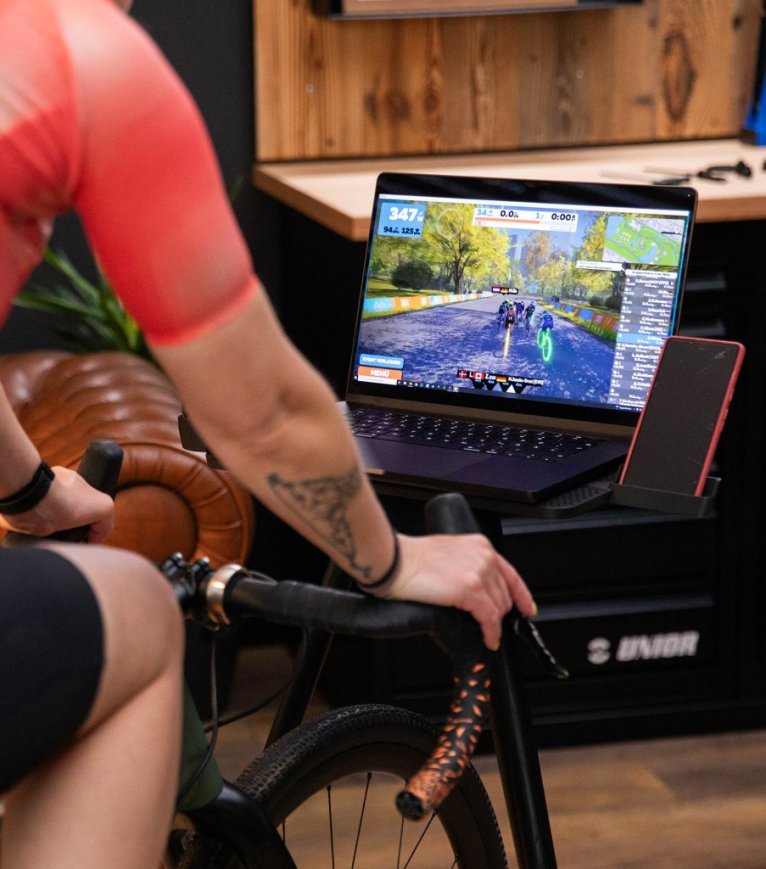 Gros plan sur une jeune femme qui pédale sur un vélo d'appartement dans son salon, tout en suivant son entraînement Zwift sur son ordinateur portable et en se concentrant sur le parcours virtuel.