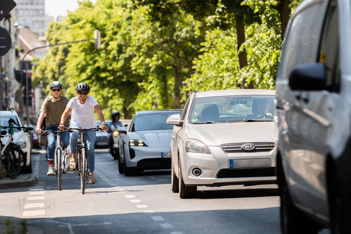 Zwei Personen fahren mit Fahrrädern auf einem Radweg in einer Stadtstraße, während Autos im Stau daneben stehen. Im Hintergrund sind Bäume und Gebäude zu sehen.