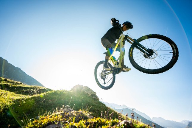 A man on an E-MTB jumps a big feature – the bike seems to hover in the air above the camera, blue sky in the background