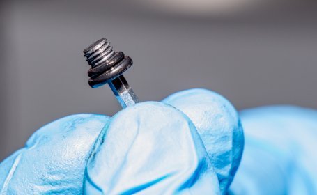 Eine Hand mit blauen Handschuhen hält eine Entlüftungsschraube mit einer schwarzen Dichtung, um sie zu überprüfen, mit unscharfem Hintergrund.