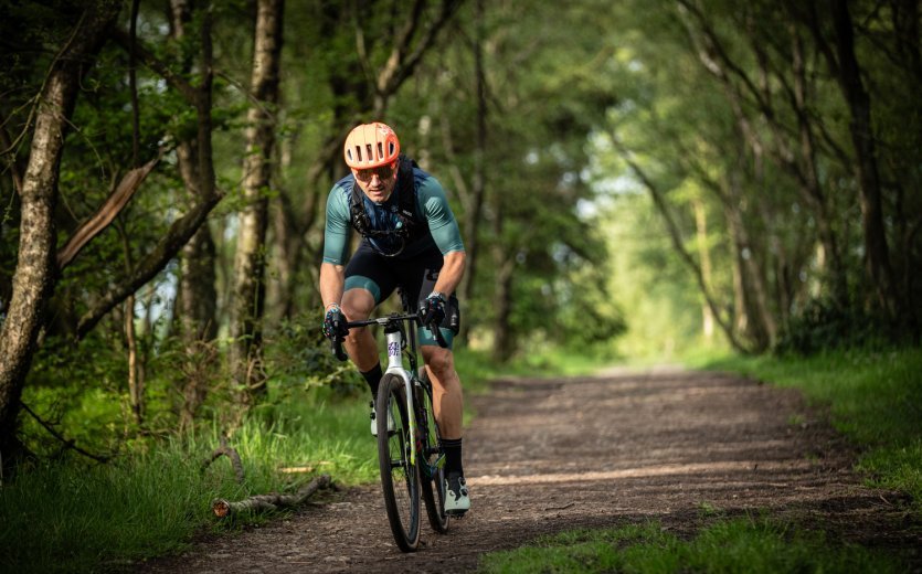 Cyclist riding a gravel bike on a forest trail, hands resting on the brake/shift levers (STI) in a slightly upright riding position, wearing helmet and cycling kit.