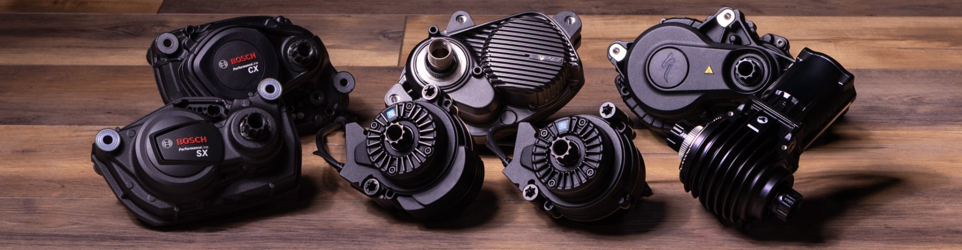 Seven different black e-bike drive units lie on a dark wooden surface.
