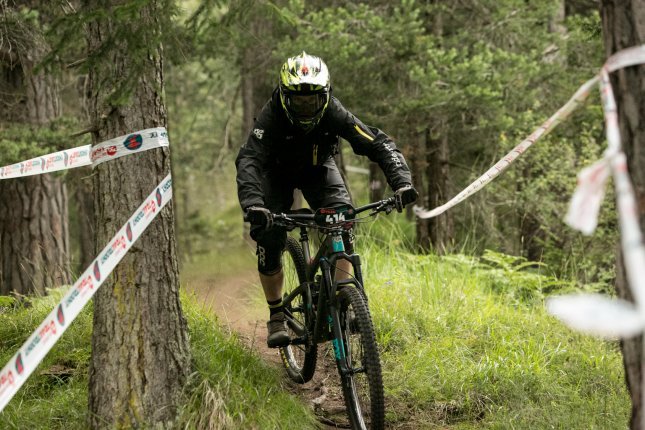 A mountain biker in black protective gear and a helmet with a visor rides on a narrow forest trail. The terrain is muddy and lined with trees. Barrier tapes along the path suggest a race or a marked course.