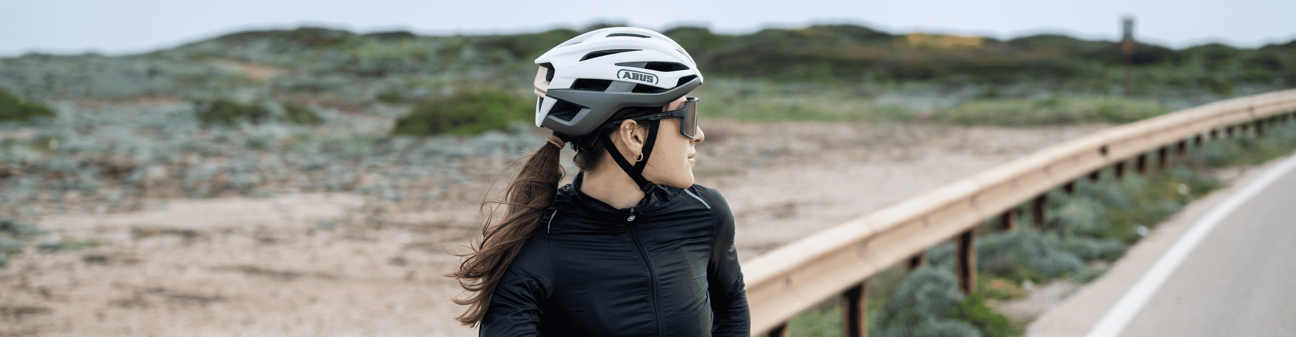 Rennradfahrerin mit ABUS Fahrradhelm steht mit ihrem Rennrad am Rand einer Küstenstraße.
