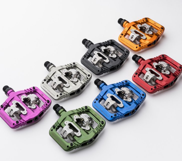 Seven colorful OneUp Clip Pedals for MTB lie side by side on a white surface.