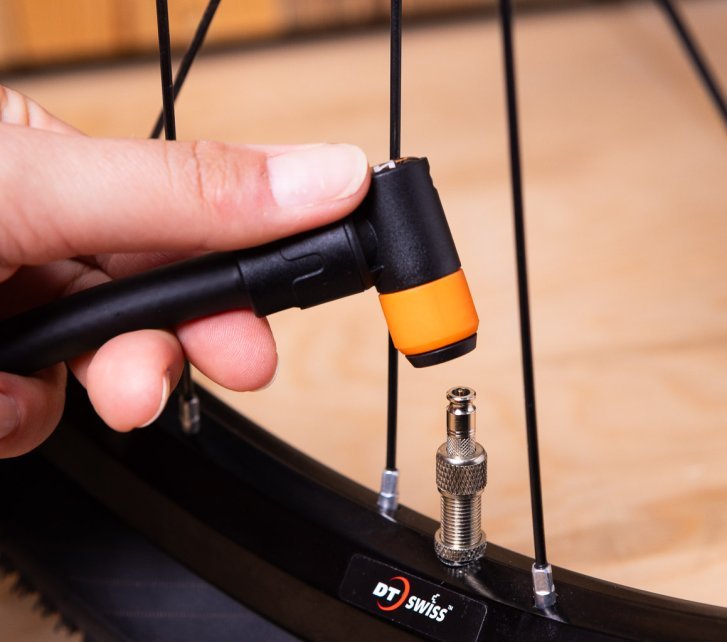 Close-up: A hand holds a black Clik Valve pump head with an orange ring next to a mounted Clik Valve.