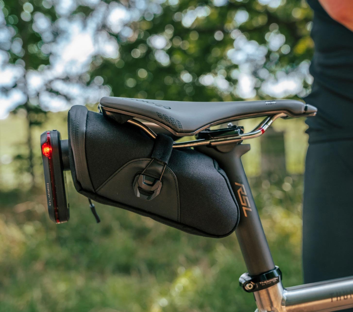 KOM Cycling saddle bag mounted beneath a road or gravel bike saddle with rear light, photographed outdoors in the forest