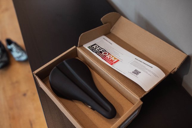 Reform-Fahrradsattel in geöffneter Versandverpackung mit beiliegender Bedienungsanleitung auf einem Tisch.