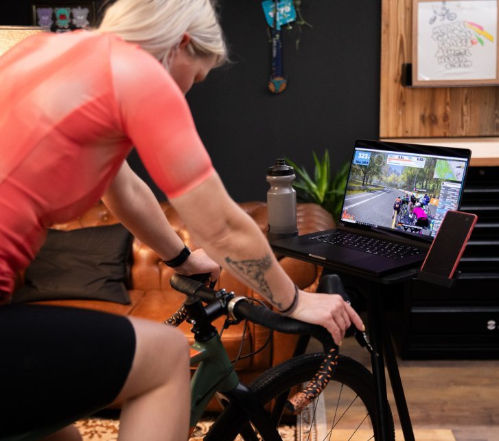 Una mujer rubia entrena en un rodillo en un taller acogedor y bien equipado. Frente a ella hay una consola con una tablet, una botella y un smartphone.