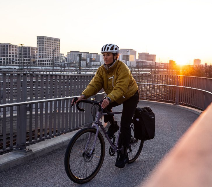 Eine Frau auf einem Rennrad fährt über eine Bahnüberführung. Dahinter sehen wir Bürohäuser und Bahngleise im Gegenlicht der untergehenden Sonne. Die Frau trägt einen weißen Helm, eine ockerfarbene warme Jacke hinten am Rad ist eine schwarze Panniertasche.