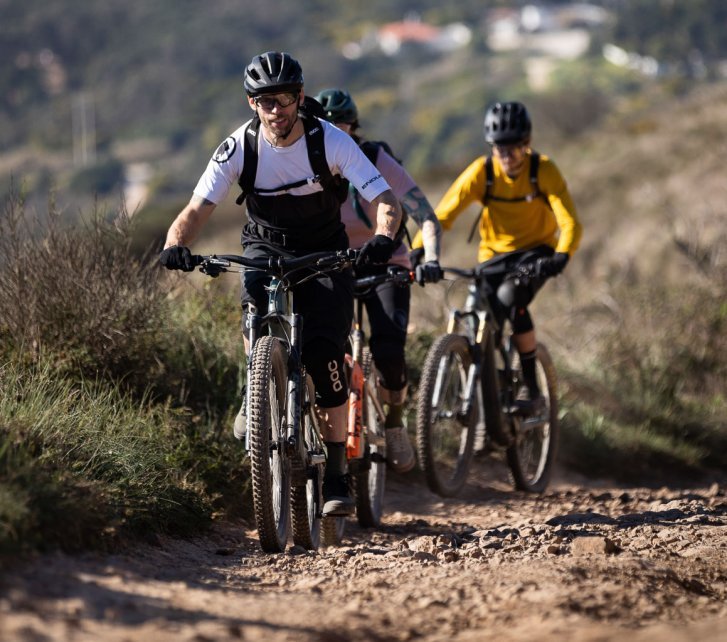 Drei E-Mountainbiker fahren auf die Kamera zu. Der Trail ist felsig und scheint leicht bergauf zu führen. Sonnenlicht wirft klare Schatten, im Hintergrund ist eine mediterrane, hügelige Landschaft zu erkennen.