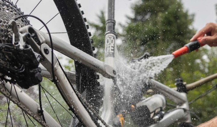 Mann mit Helm reinigt ein Mountainbike im Freien mit einem Wasserschlauch. Das Hinterrad ist angehoben, Wasser spritzt auf Rahmen, Antrieb und Kassette. Das Fahrrad wird gründlich abgespült, um Schmutz und Rückstände zu entfernen.