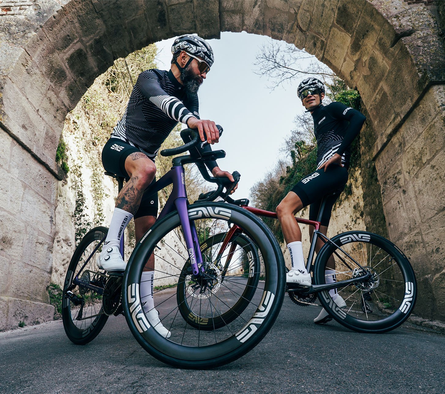 Dos ciclistas de carretera con bicicletas ENVE de alto rendimiento y ruedas aerodinámicas bajo un arco histórico de piedra en una carretera tranquila. Perspectiva dinámica centrada en componentes de carbono premium.