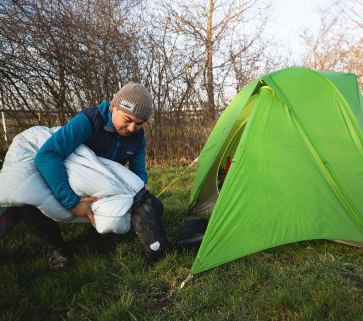 Marcel from bc Marketing stuffs his sleeping bag into its stuff sack. He is kneeling in front of the entrance of his tent.