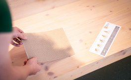 Two hands place a rectangular piece of corrugated cardboard on a light wooden bench. Next to it lies a measuring gauge from SQlab.