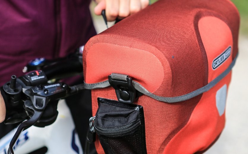 A red Ortlieb bicycle bag is attached to a bike. In the foreground, part of the handlebar with shifters and a hand are visible, while a blurry green field can be seen in the background.