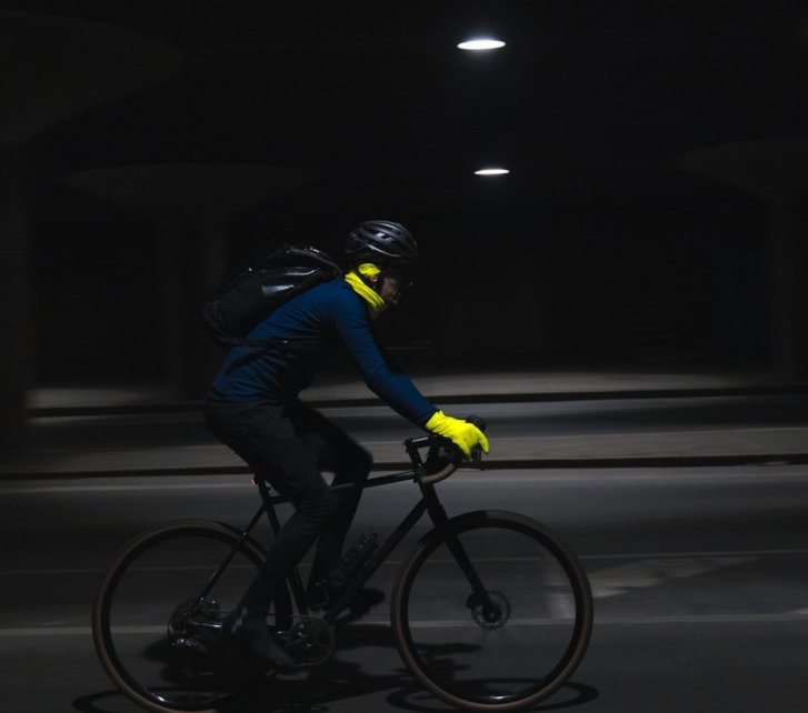 Eine Person auf einem dunklen Rennrad fährt quer zur Blickrichtung nach rechts. In der nächtlichen Szenerie sind lediglich die neongelben Handschuhe und ein neongelbes Halstuch klar erkennbar.