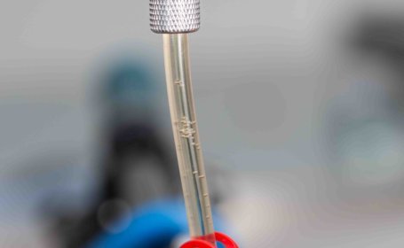 In einem transparenten Schlauch steigen kleine Luftblasen durch eine klare gelbe Flüssigkeit in eine Spritze auf. Ein roter, offener Schlauchklemmring und ein metallischer Adapter zum Bremszylinder sind sichtbar.