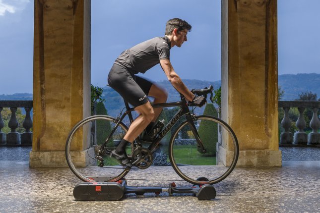 A man trains on a road bike set up on free rollers in front of a Mediterranean backdrop.
