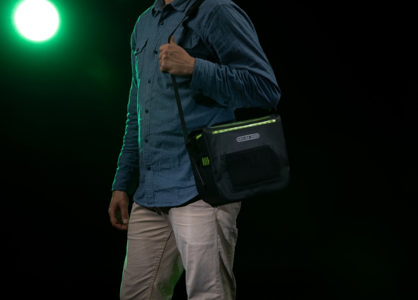 Eine Person mit einer schwarzen Fahrradtasche mit grünen Akzenten über der Schulter steht vor einem grünen Licht. Die Tasche hat ein beleuchtetes Design. Der Hintergrund ist dunkel.