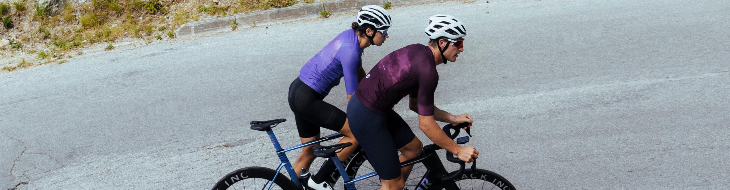 Zwei Rennradfahrer in Castelli Radsportbekleidung fahren bergauf auf einer Asphaltstraße, Aero-Laufräder und Helme.