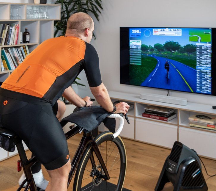 Un hombre con un maillot naranja entrena en un rodillo en un salón. Frente a él, la plataforma de entrenamiento Zwift se muestra en un televisor.