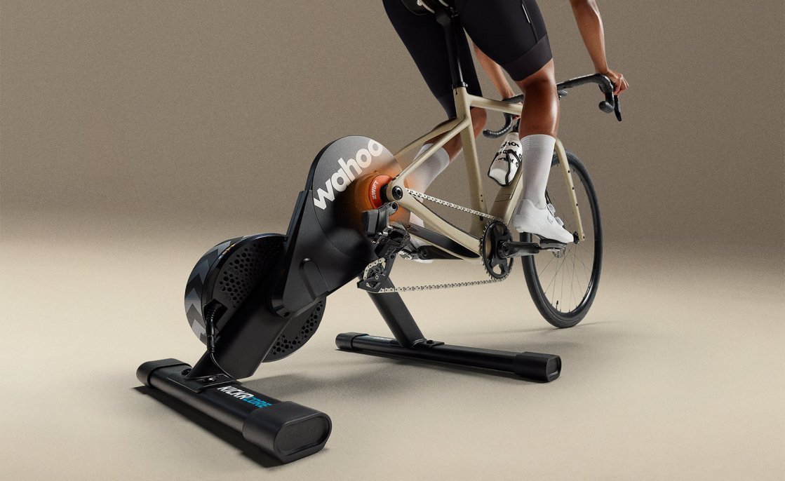 Un vélo de course est monté sur le home trainer Wahoo KICKR Core avec Zwift Click & Cog, tandis qu'une personne s'entraîne en salle dans un environnement de studio.