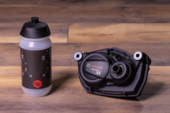 A Bosch SX e-bike drive lies on a dark wooden floor. A bike water bottle stands next to it