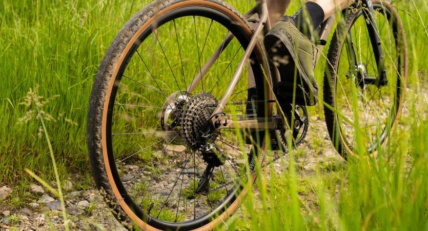 Roue arrière d’un vélo gravel sur chemin en gravier, pneu à flanc beige en action, cassette et dérailleur visibles dans un décor verdoyant.