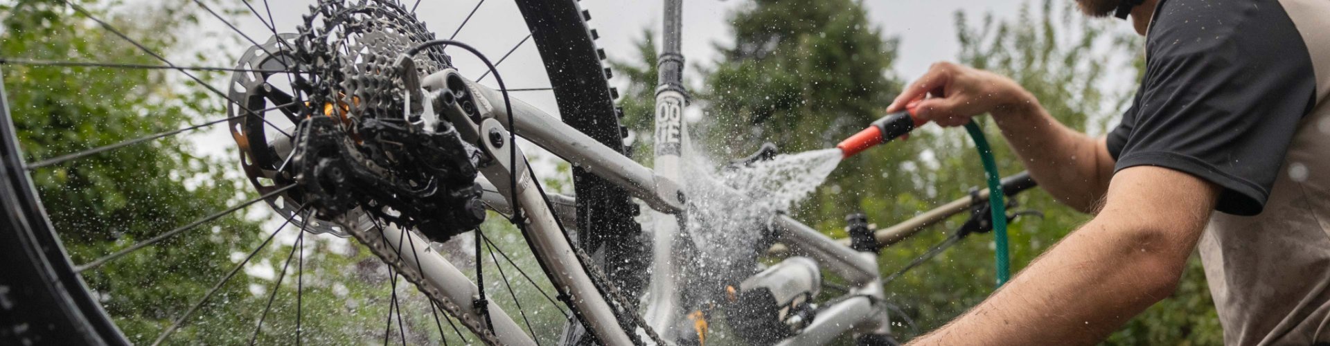 Mann mit Helm reinigt ein Mountainbike im Freien mit einem Wasserschlauch. Das Hinterrad ist angehoben, Wasser spritzt auf Rahmen, Antrieb und Kassette. Das Fahrrad wird gründlich abgespült, um Schmutz und Rückstände zu entfernen.
