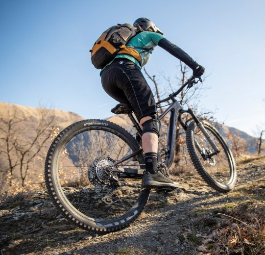 Eine Mountainbikerin fährt auf steinigem Gelände bergauf. Sie trägt einen Rucksack und Bike Wear passend zu dem frühlingshaften Wetter.