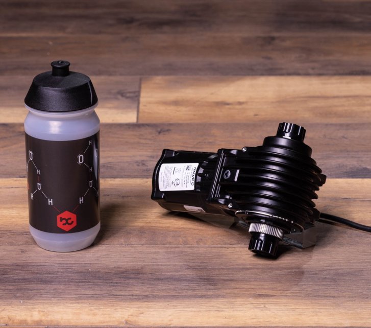 A Fazua Ride 60 e-bike drive lies on a dark wooden floor. A bike water bottle stands next to it